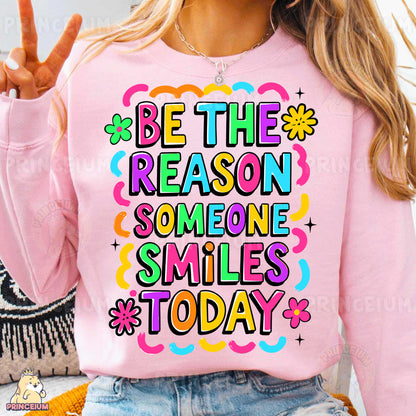 a woman wearing a pink sweatshirt that says be the reason someone smiles today